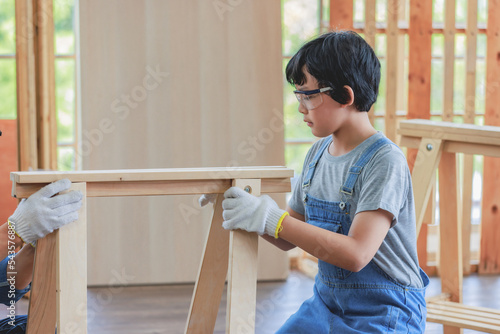 Asian amateur young boy in jeans outfit with safety glasses goggles and gloves helping unrecognizable unknown father holding assembling wooden furniture in home housing building construction site
