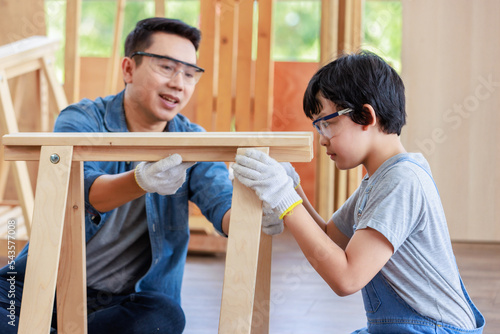 Asian amateur young boy in jeans outfit with safety glasses goggles and gloves helping professional carpenter engineer father holding assembling wooden furniture in housing building construction site