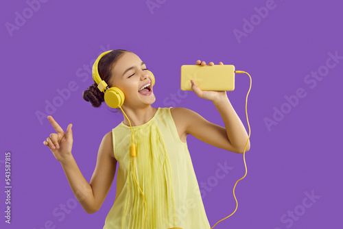 Cheerful preteen girl in headphones connected to mobile phone listens to music and sings along. Funny kid girl in stereo headphones uses phone as microphone having fun on purple background. Banner.