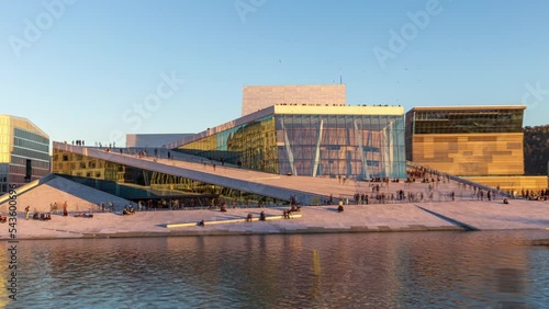 Oslo opera house hyper-lapse with people walking in the afternoon light, on a sunny day under clear blue sky. Time-lapse of the famous landmark in the capital of Norway, with water in the foreground.