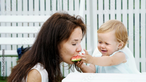 summer, in the garden, a funny one-year-old blonde girl treats her mother with watermelon, feeds her from her hands, the girl also eats watermelon. High quality photo