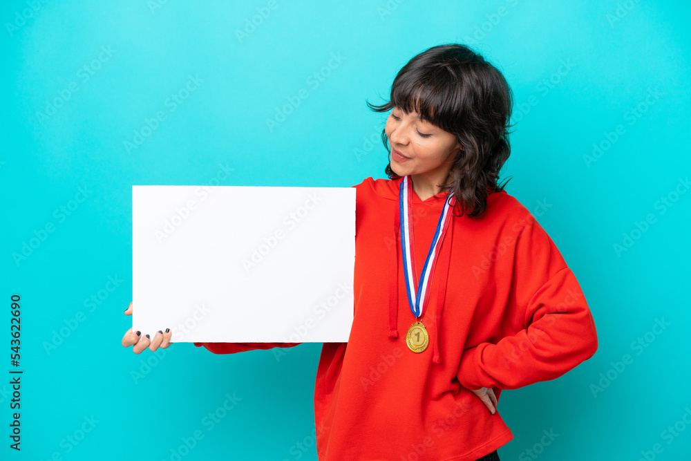 Young latin woman with medals isolated on blue background holding an empty placard with happy expression and pointing it Young latin woman with medals isolated on blue background holding an empty placard with happy expression and pointing it