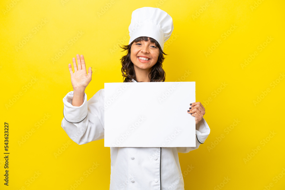 Young chef latin woman isolated on yellow background holding an empty placard and saluting Young chef latin woman isolated on yellow background holding an empty placard and saluting