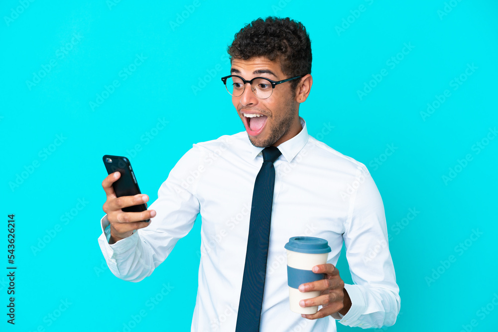 Young business Brazilian man isolated on blue background holding coffee to take away and a mobile Young business Brazilian man isolated on blue background holding coffee to take away and a mobile