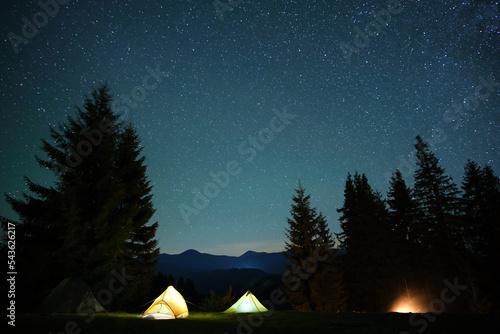 Bright illuminated tourist tents near glowing bonfire on camping site in dark mountain woods under night sky with sparkling stars. Active lifestyle and outdoor living concept © bilanol