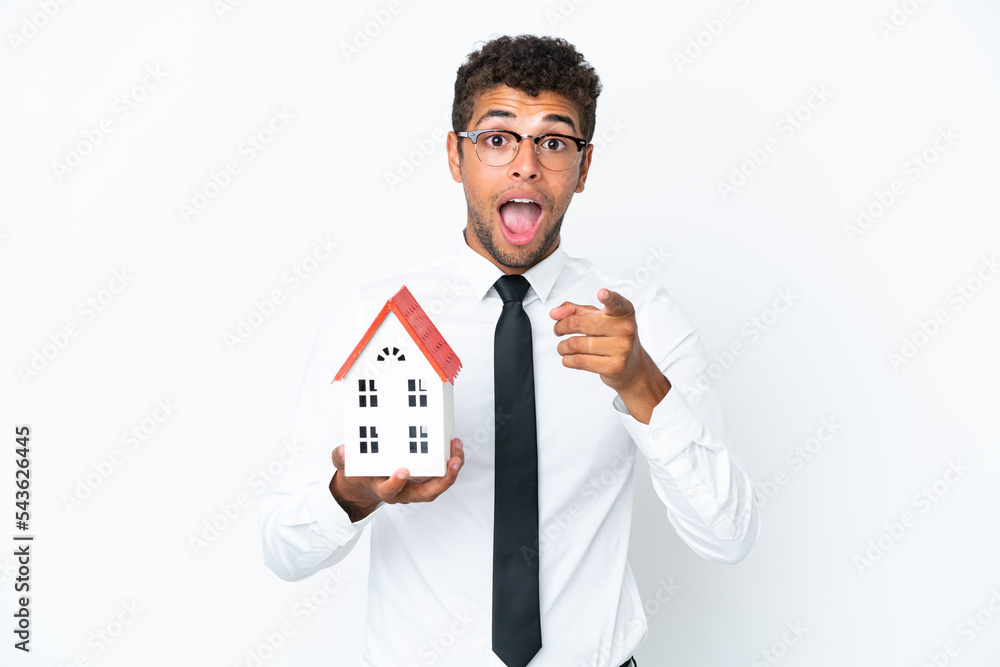 Young business Brazilian man holding a house toy isolated on white background surprised and pointing front Young business Brazilian man holding a house toy isolated on white background surprised and pointing front