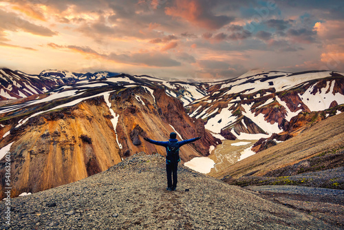 Hiker man standing with arms raised on the cliff with volcanic mountain and snow covered from Blanhjukur trail on summer in the evening photo