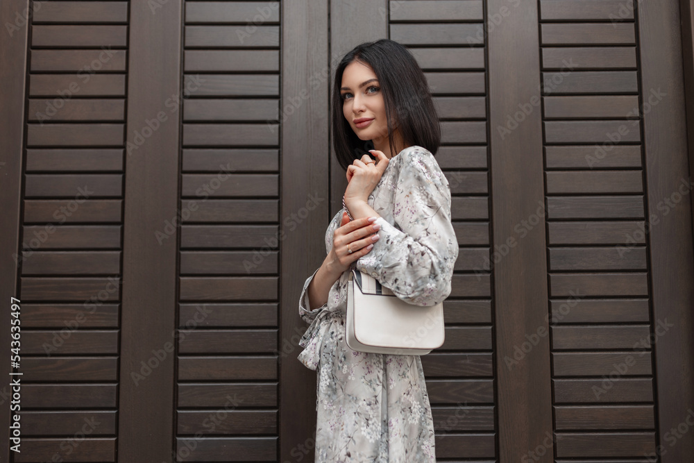 Fototapeta premium Fashionable beautiful girl in a stylish vintage summer dress with a floral print with a white trendy handbag stands near a brown wooden door