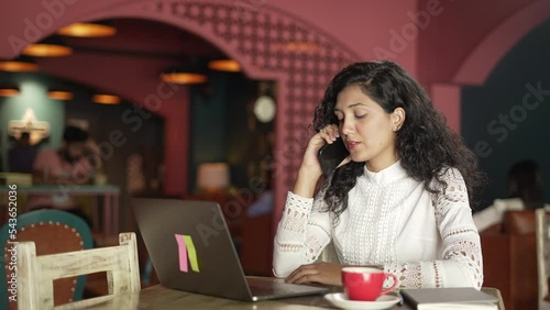  young Indian business woman using laptop talking on the phone having in cafe shop. smiling Asian  girl siting on chair in the laptop.smartphone talks to client distantly, provide help by phone call. photo