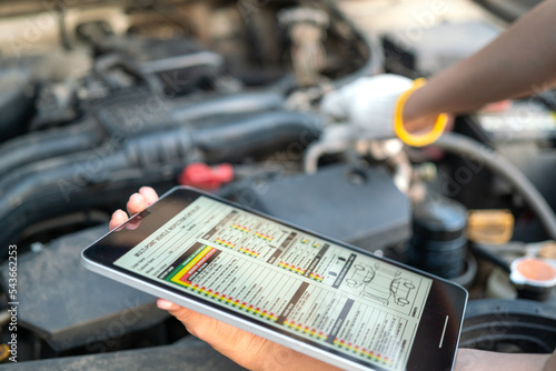 фотографія Using the vehicle checklist guideline on the digital tablet during perform preventive maintenance with car engine part as blurred background