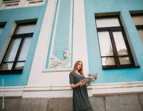 A young pretty girl with blond hair, in a gray dress with a paper guide in her hands, stands near the blue facade of an ancient museum, studies the travel route. photo