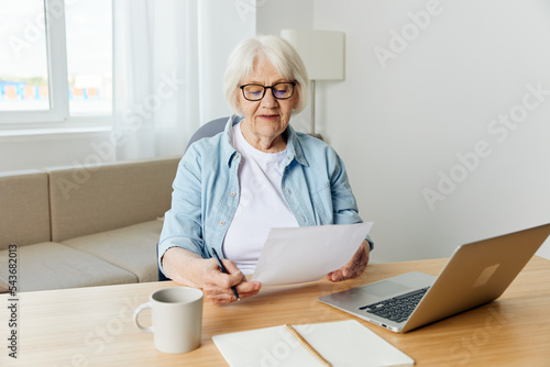 Senior white haired woman wearing glasses. sitting with laptop at the table in her comfortable flat and happily smiling works