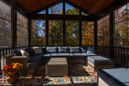 Cozy screened porch enclosure with contemporary furniture at Thanksgiving Holiday. Flower bouquet in a vase, autumn leaves and woods in the background.