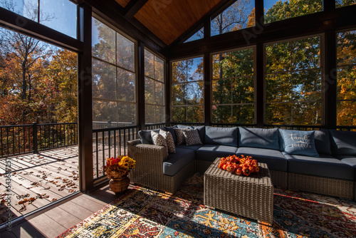 Cozy screened porch enclosure with contemporary furniture at Thanksgiving Holiday. Porch door open, flower bouquet in a vase, autumn leaves and woods in the background.