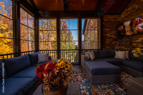 Cozy screened porch enclosure with contemporary furniture at Thanksgiving Holiday. Flower bouquet in a vase, autumn leaves and woods in the background.