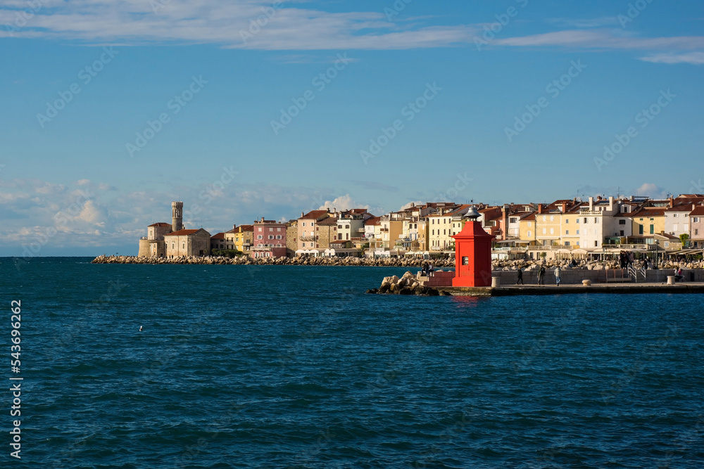 A small red lighthouse on the waterfront of the historic medieval town of Piran on the coast of Slovenia. Background left is the Our Lady of Health Church and a 17th century lighthouse tower

