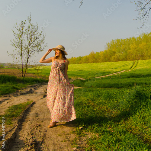 Smiling young woman in dress and hat walking down the footpath in the countryside nature on a sunny day 