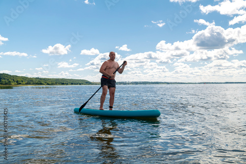 A man in shorts and a T-shirt on a SUP board in a lake on a sunny day against a sky with clouds. photo