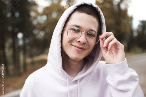 Smiling cute teenager boy 19-20 year old wear glasses and hood over nature background outdoor close up. Happy male kid student. photo