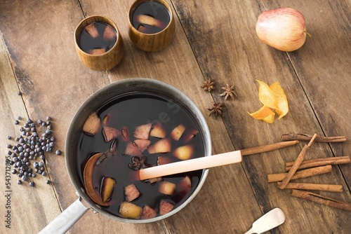 Top view of a pot of hot spicy mulled red wine surrounded by ingredients and fruit on a wooden table