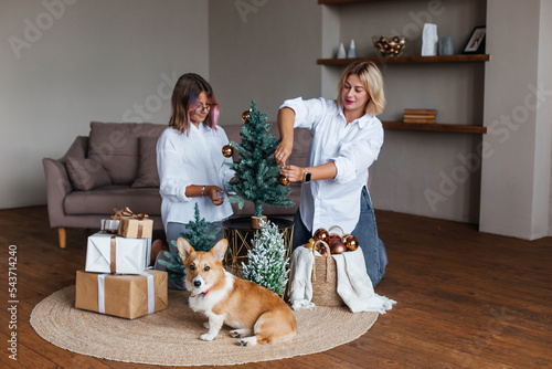 Mother and daughter near the Christmas tree with a corgi dog. Mom and daughter decorate the Christmas tree together with Christmas balls, play with the dog, have fun © Наталья Мокрецова