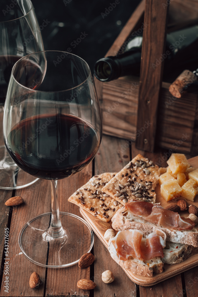 Two glasses of red wine stand on a wooden table. Prosciutto with cheese, red wine, baguette, nuts. Red wine with traditional Mediterranean snacks.