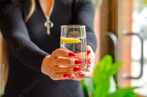 Young woman in black dress and with red manicure in cafe, holds glass of clear water with lemon. Concept is not to forget to drink clean water every day for the health of body