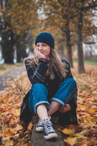 Smiling and happy brunette sitting in a pile of colourful leaves in a city park. Candid portrait of a young real woman in autumn clothes in the fall season. Orange, red and brown colour