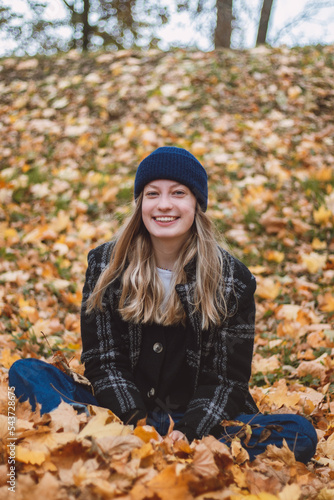 Smiling brunette sitting in a pile of colourful leaves in an oak forest throwing leaves. Candid portrait of a young real woman in autumn clothes in the autumn season