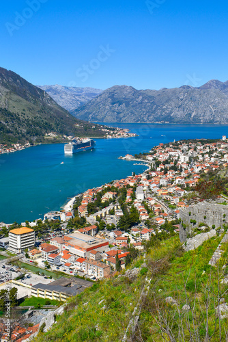 Kotor, Montenegro, Europe. Bay of Kotor on Adriatic Sea. Rock, historical walls, roofs of the buildings in the old town. Cruise in the bay, mountains in the background. Clear blue sky, sunny day.
