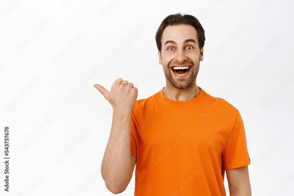 Close up of enthusiastic adult man pointing left, smiling amazed, showing advertisement aside, wearing orange t-shirt, white background Close up of enthusiastic adult man pointing left, smiling amazed, showing advertisement aside, wearing orange t-shirt, white background
