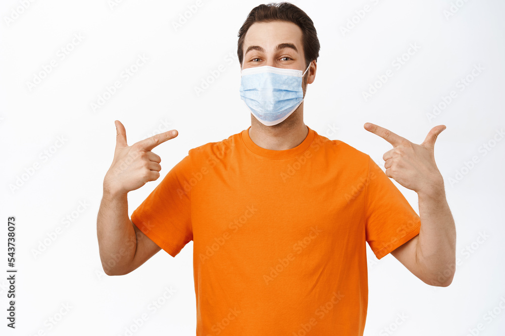 Image of smiling man points at his face with medical face mask, wears orange t-shirt, white background Image of smiling man points at his face with medical face mask, wears orange t-shirt, white background