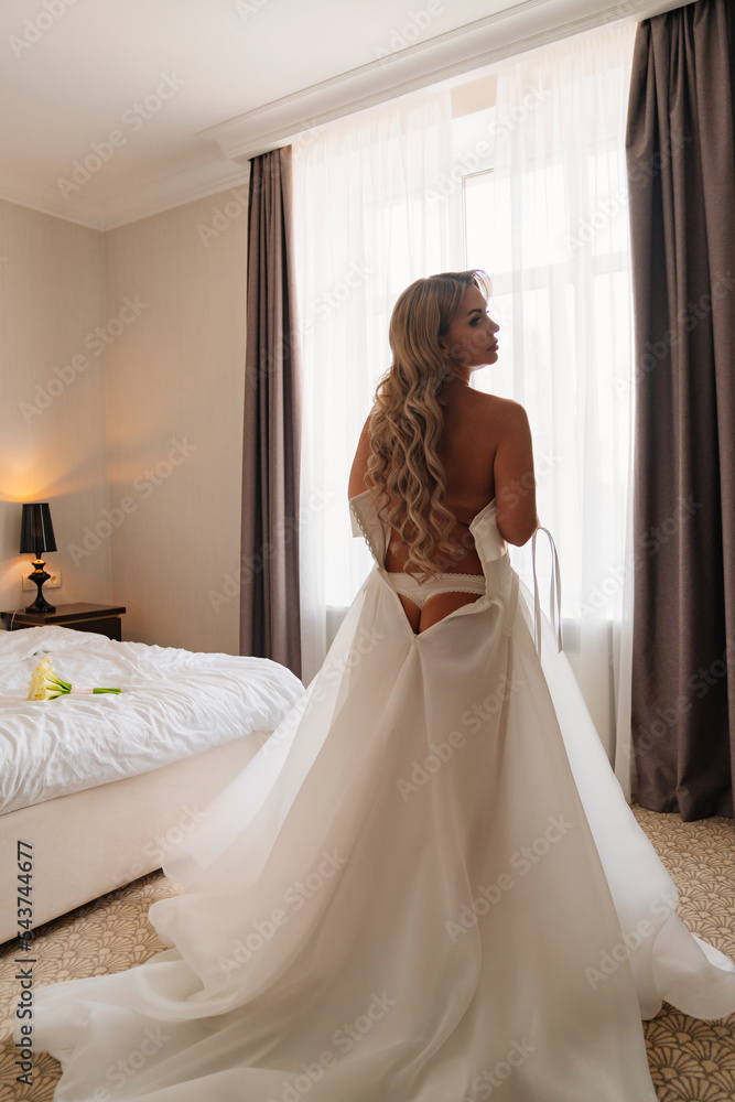 Fototapeta premium view from the back. a blonde bride wears a white dress by the bedroom window.