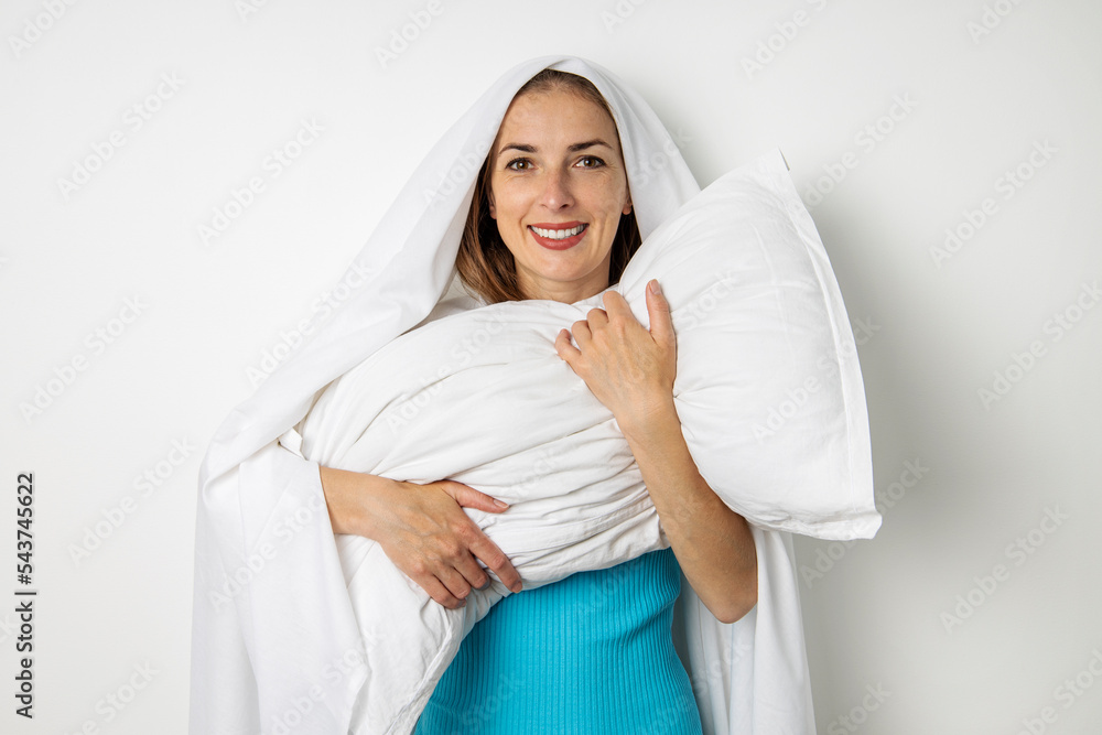 Smiling young woman hiding herself in a sheet hugging a pillow on a white background