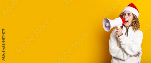 Young woman in santa claus hat shouts into a shout on a yellow background. Banner.
