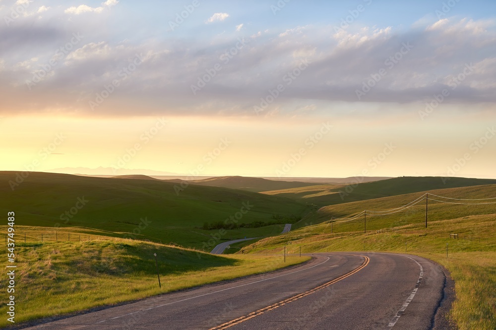 A simple image with a road leading into a hilly landscape. It is sunset with a beautiful glow and clouds in the sky catching the yellow light. Landscape with nobody in the image.