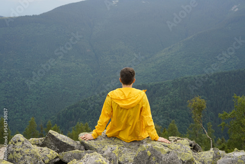 A young man in a yellow raincoat sits on a rock and enjoys the view high in the mountains. The concept of ecological tourism and healthy recreation