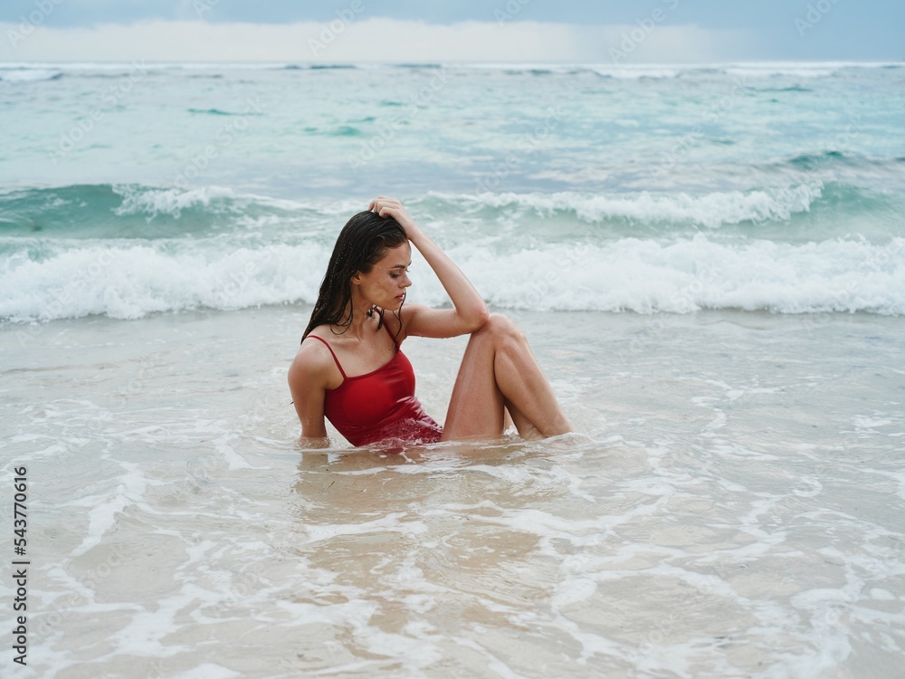 Woman with a beautiful tan tourist in a red swimsuit sitting on the sand on the beach in the ocean in the waves, cloudy weather Woman with a beautiful tan tourist in a red swimsuit sitting on the sand on the beach in the ocean in the waves, cloudy weather