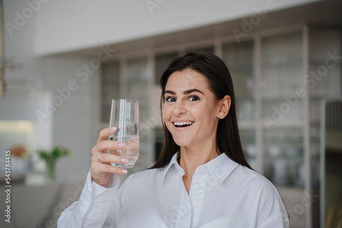 Amazed brunette housewife smiles wide looks at camera holds glass of water dressed in white shirt against blurry living room. Healthy lifestyle and skincare. Cute hispanic female enjoys pure water.