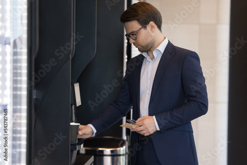 Young businessman in suit pours favourite beverage, uses vending machine in office cafeteria or self-service automated retail place for comfort and busy life, start working day with cup of hot coffee photo