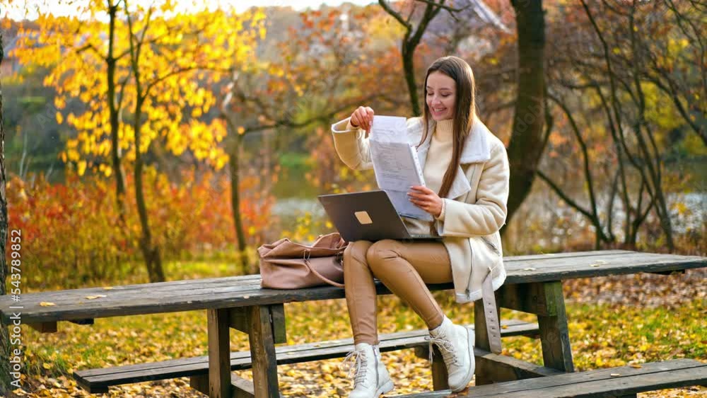 Happy woman working remotely in the autumn park. Lady checking up the documents and holding computer on her laps. Blurred backdrop. Happy woman working remotely in the autumn park. Lady checking up the documents and holding computer on her laps. Blurred backdrop.