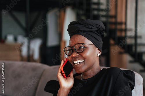 Fotografia Close up of African young Woman in Black turban, glasses toothy smiling talks by phone sitting at home on cozy sofa