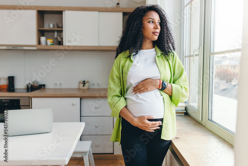 Indoor portrait of african american pensive pregnant woman in green shirt, waiting for her husband to come back from business trip, standing in kitchen looking through window, rubbing cute belly