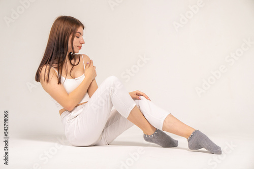 Cute young brown-haired girl in a white top and knitted trousers posing while sitting on a white background