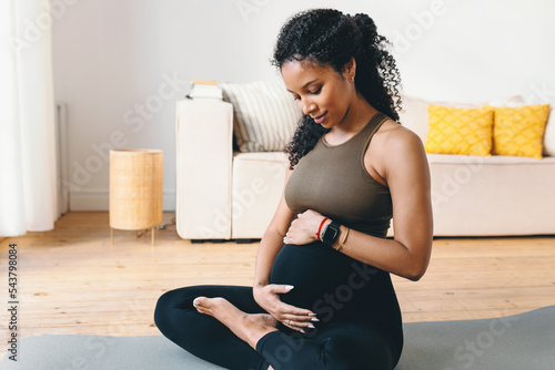 Cute pregnant female of black ethnicity sitting on mat in sports clothes rubbing her adorable belly, having rest in fitness workout at home, wearing smart watch to control pulse and heart rate