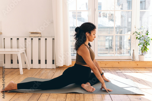 Prenatal fitness training. Sporty african american pregnant woman in leggings sitting on left leg, stretching right, practicing prenatal yoga or pilates on mat at home against window photo