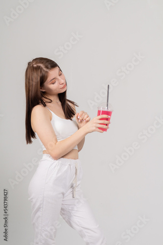 A cute young brown-haired girl drinks a cocktail from a glass with a straw on a white background The model is dressed in a white top and knitted trousers