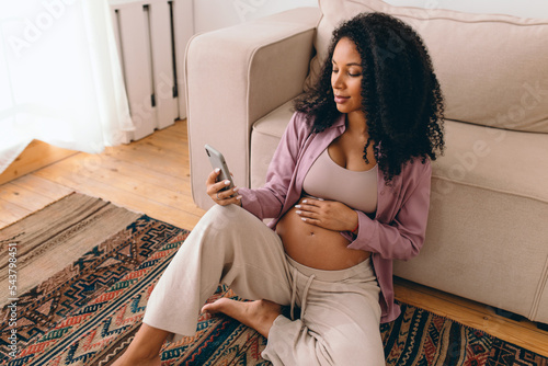 Portrait of gorgeous pregnant female with dark skin and curls sitting on carpet next to couch browsing news feed in her social media app, reading article, watching podcast for future moms