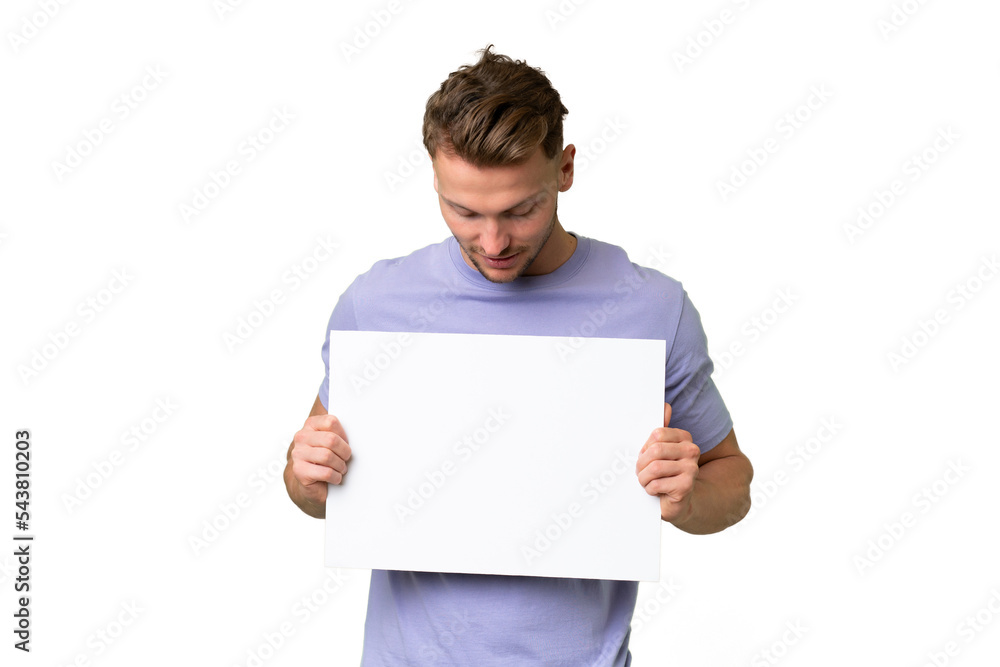 Young blonde caucasian man over isolated background holding an empty placard and looking it Young blonde caucasian man over isolated background holding an empty placard and looking it