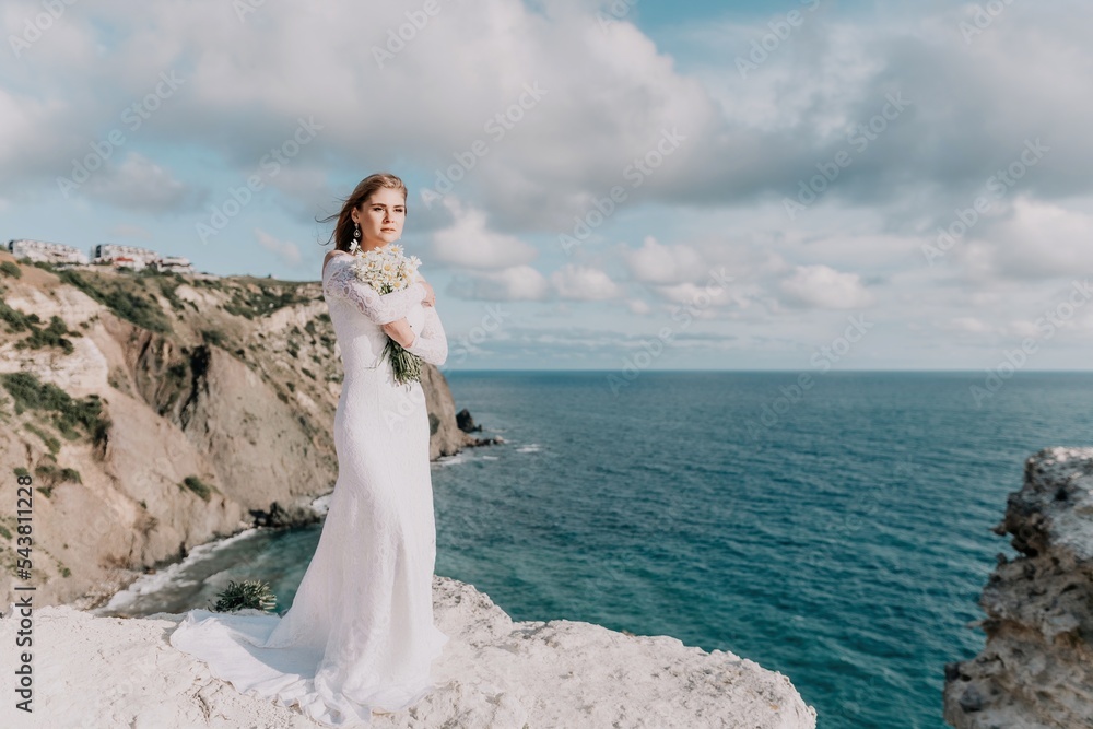 Romantic bride, blond girl in white wedding dress with open shoulders posing with sea and rocks in background. Stylish young woman standing on cliff's edge and looking into a ocean mountains at sunset Romantic bride, blond girl in white wedding dress with open shoulders posing with sea and rocks in background. Stylish young woman standing on cliff's edge and looking into a ocean mountains at sunset